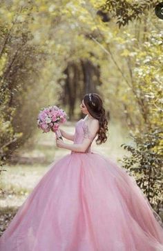 a girl in a pink dress holding flowers and looking at the ground with trees behind her