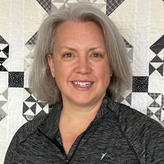 an older woman with grey hair smiles in front of a quilted wall and floor