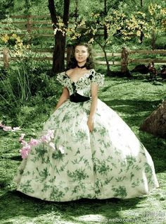 a woman in a long dress standing next to a tree with flowers on the grass