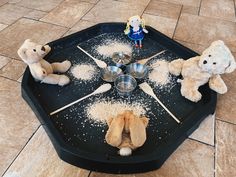 teddy bears sitting around a black tray with food on it and measuring spoons in the middle