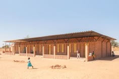 two people are walking in front of a small building on dirt ground with several doors and windows