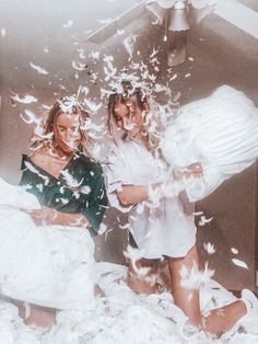 two women are in bed with white sheets and feathers flying around them as they look at the camera