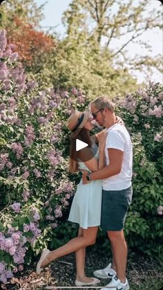 a man and woman standing next to each other in front of some bushes with purple flowers