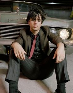 a man sitting in front of a car wearing a suit and tie with his hands on his hips