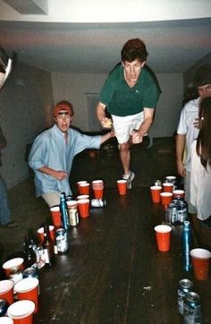 a group of people standing around a wooden table with cups on it and one person reaching for the cup