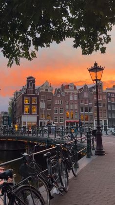 bicycles are parked on the side of a bridge near water and buildings with lights at sunset