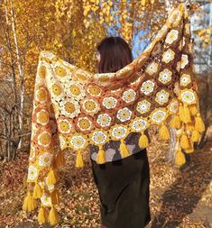 a woman is walking in the woods carrying a crocheted shawl with tassels