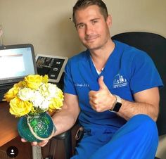 a man in blue scrubs is holding a vase with yellow flowers and giving the thumbs up