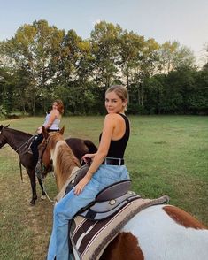 two women riding on the backs of brown and white horses in a grassy field with trees behind them