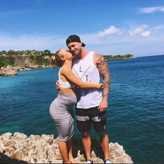 a man and woman standing next to each other on top of a cliff near the ocean