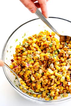 a person holding a spoon in a bowl filled with corn and other food on the table