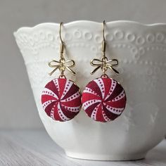 "These festive peppermint earrings will add a delightful touch of holiday magic to your look. They're comfortable for everyday wear or for special occasions like to a winter dinner party or on Christmas Day. The earrings are handmade with swirled red and white peppermint fabric, brass backings, and cute gold bows. The hooks are made of hypoallergenic gold stainless steel, and they come with clear rubber backings so they will stay on easier. At just 3/4\" wide, these earrings are not tiny yet not large, making them and ideal size for just about anyone! They measure about 1 5/8\" long (that's a little over one and a half inches).  Details: Full length - about 1 5/8\" Width - 3/4\"  Earwire - hypoallergenic gold stainless steel Earring backing - clear rubber Gift boxes: All jewelry comes in a Handmade Christmas Earrings For Celebration, Handmade Christmas Celebration Earrings, Red Round Earrings For Christmas, Red Dangle Earrings For Holiday, Red Drop Earrings For Holiday, Handmade Round Christmas Earrings, Red Dangle Earrings For Christmas, Festive Christmas Earrings, Festive Christmas Round Earrings