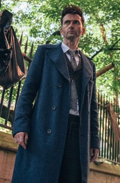 a man wearing a coat and tie standing in front of a fence with a bag on it