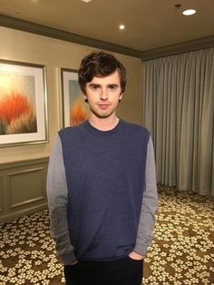 a young man standing in front of a painting on the wall and wearing a blue sweater