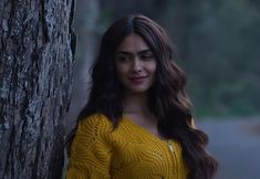 a woman standing next to a tree with long hair and wearing a yellow sweater in the woods