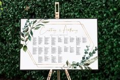 a wedding seating sign with greenery and gold foil on it, sitting in front of a hedge