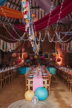 This shows a colourful wedding set up with three rectangular rows of dining tables. The focus is on the middle row of wooden tables. On the table is a pink table runner across the middle of the table. There are also black plates paired with many different coloured napkins. On the table are glassware and glass vases with flowers in them. There are also brass candlesticks with colourful candles.Above the rows of tables are colourful bunting. At the base of the table are round paper lanterns. Lake Party, Paper Lanterns Wedding, Wedding Setup, Wedding Dining, Dinner Setting, Vibrant Wedding, Picnic Wedding, Wedding Venue Decorations, Wedding Set Up