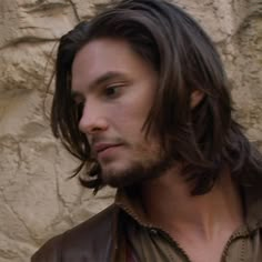 a man with long hair wearing a leather jacket and looking off to the side while standing in front of a stone wall