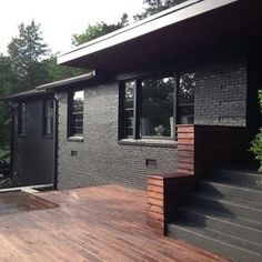 a wooden deck with steps leading up to a black brick house in the background, surrounded by greenery and trees