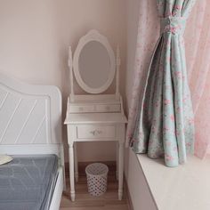 a bedroom with a bed, desk and mirror next to a window covered in pink curtains