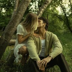 a man and woman are sitting in the grass by some trees, one is kissing the other's cheek