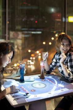 two women sitting at a table with drinks in front of them and one woman holding up her hand