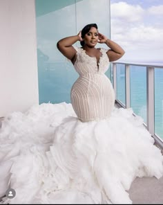 a woman in a white gown standing on a balcony next to the ocean with her hands behind her head