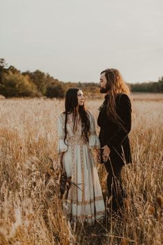 two women standing next to each other in a field with tall grass and trees behind them