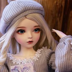 a doll with blonde hair wearing a gray knitted sweater and hat holding a card