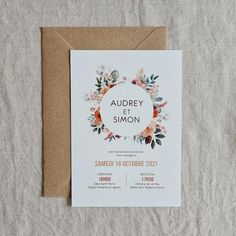 a wedding card with an orange and white floral wreath on it, next to a brown envelope