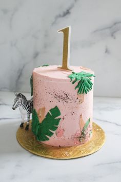 a pink cake decorated with palm leaves and a gold number one on top, sitting on a marble table