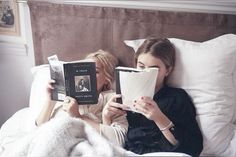 two women laying in bed reading books and looking at each other's faces with their eyes closed