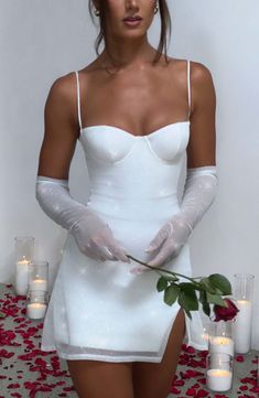 a woman in white dress and gloves holding a rose with candles on the table behind her