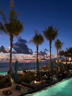 an outdoor pool with palm trees and lights on the water at dusk, next to a beach