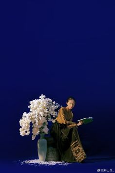 a woman is sitting next to a vase with flowers in it and holding a book