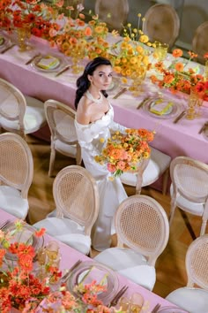 a woman in white dress standing next to table with flowers on it and chairs around her