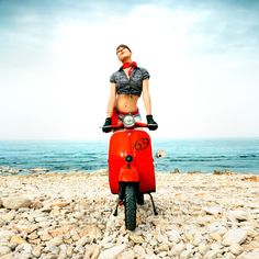 a woman sitting on top of a red scooter next to the ocean and rocks