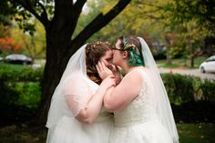 two brides kissing each other in front of a tree and grass covered park area