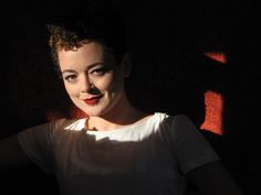 a woman with red lipstick standing in the dark, wearing a white shirt and looking at the camera