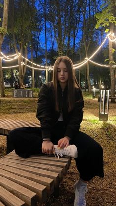 a woman sitting on a wooden bench in a park at night with string lights strung across the trees