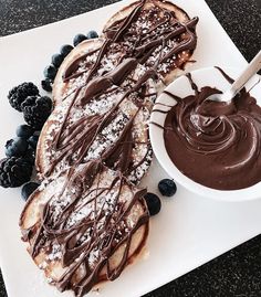 a white plate topped with pancakes covered in chocolate frosting and blueberries next to a bowl of fruit