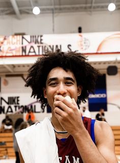 a young man with curly hair eating a hot dog in a gym while wearing a red and white shirt