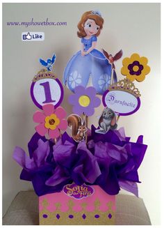 a purple and gold birthday centerpiece with princesses on it's top, surrounded by flowers