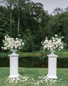 two white pedestals with flowers on them in front of a lake and forest area