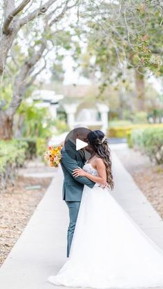 a bride and groom kissing on the sidewalk in front of some trees with their arms around each other