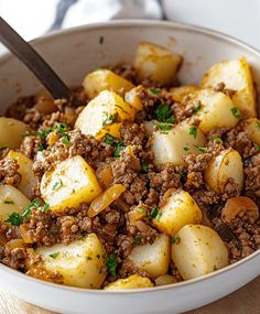 a white bowl filled with meat and potatoes on top of a wooden table next to a fork