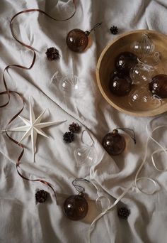 some glass ornaments are laying on a white sheet with brown ribbons and pineconis