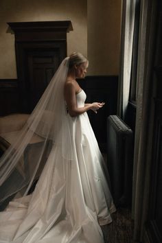 a woman in a wedding dress looking at her cell phone while standing next to a window