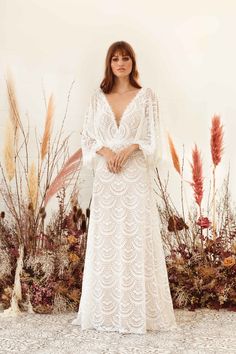 a woman standing in front of flowers wearing a white dress with long sleeves and an open back