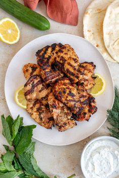 grilled chicken on a plate next to pita bread, cucumbers and lemon wedges